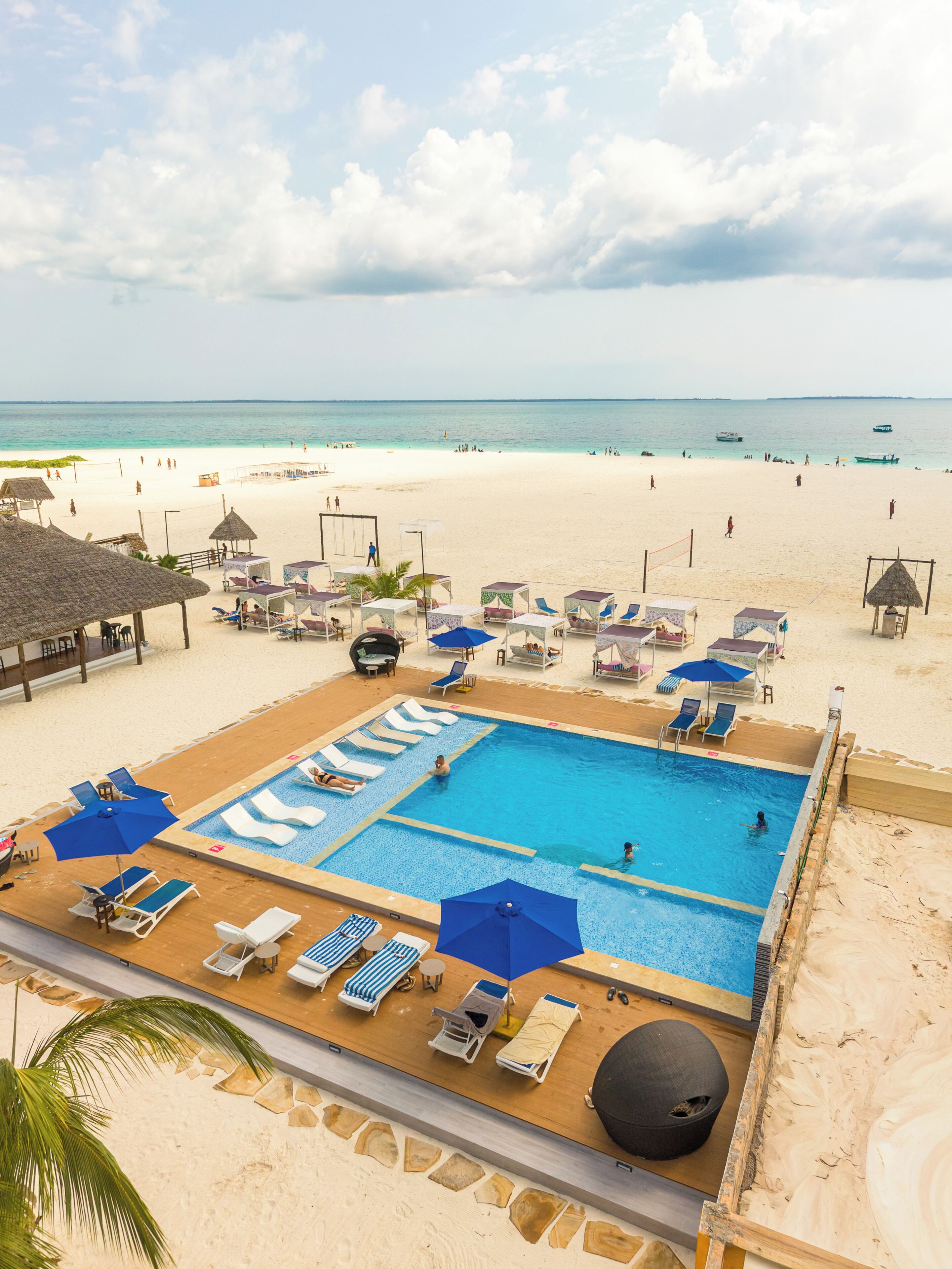 A pool area with lounge chairs and umbrellas, surrounded by a sandy beach and a clear sky.