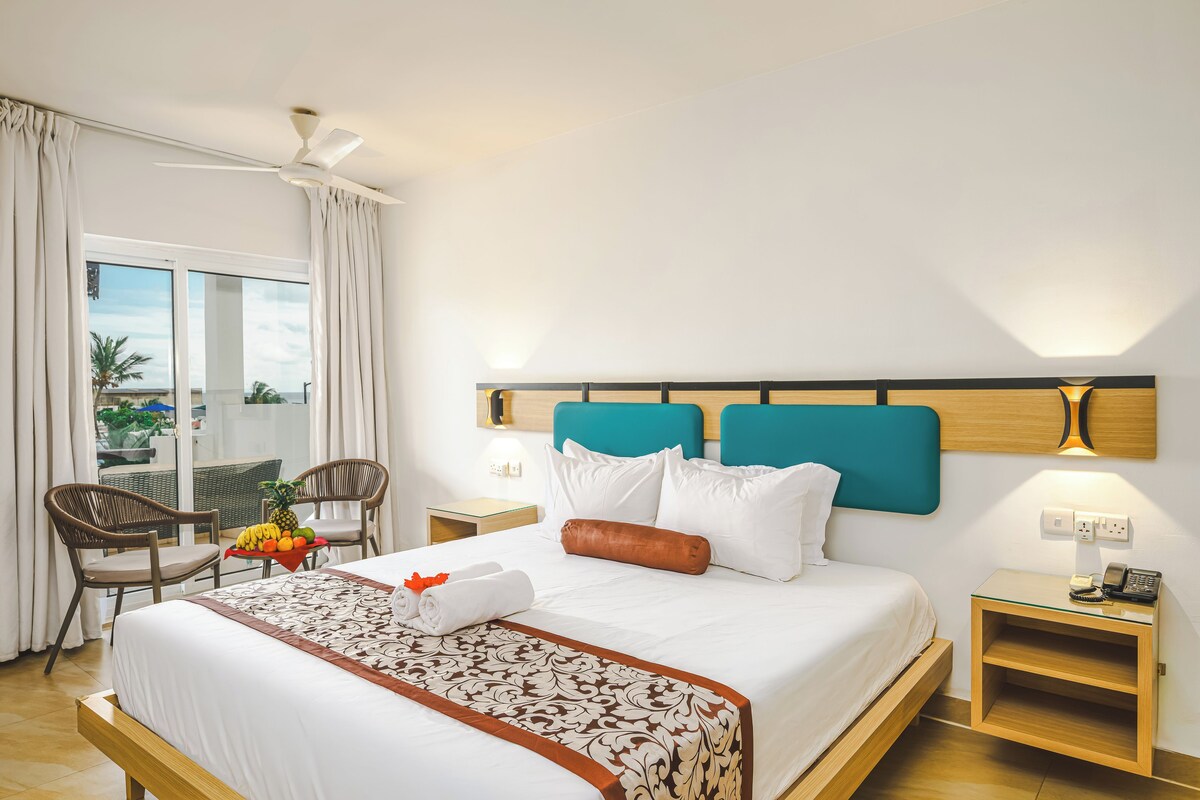 A hotel room with a bed, a chair, a table with fruit, a ceiling fan, and a view of the outdoors.