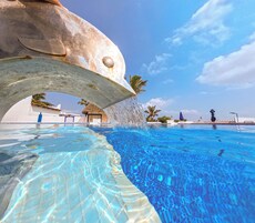 A swimming pool with clear blue water, a large fish sculpture, and palm trees in the background.