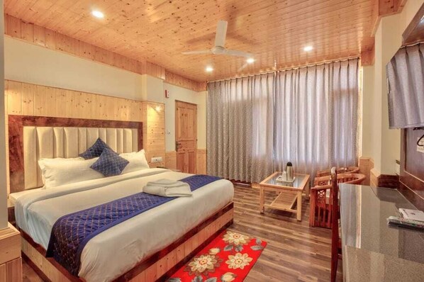 A hotel room with a wooden ceiling, a large bed with blue bedding, a small table with chairs, and a window with curtains. - OnlyStay Surya Mall Road Manali (Manali)