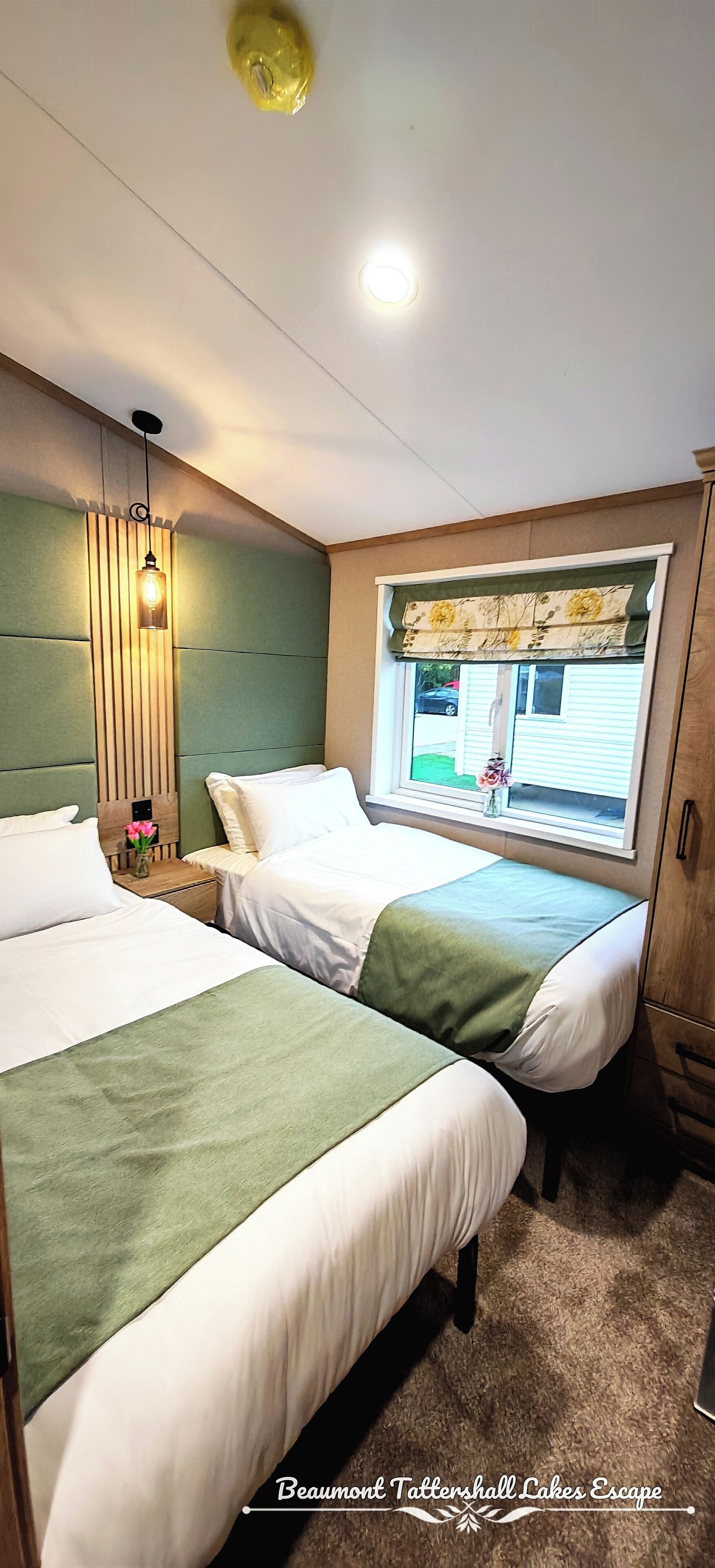 Habitación de hotel con dos camas, ventana con vistas, cabecero de madera y mesita de noche con un jarrón de flores.