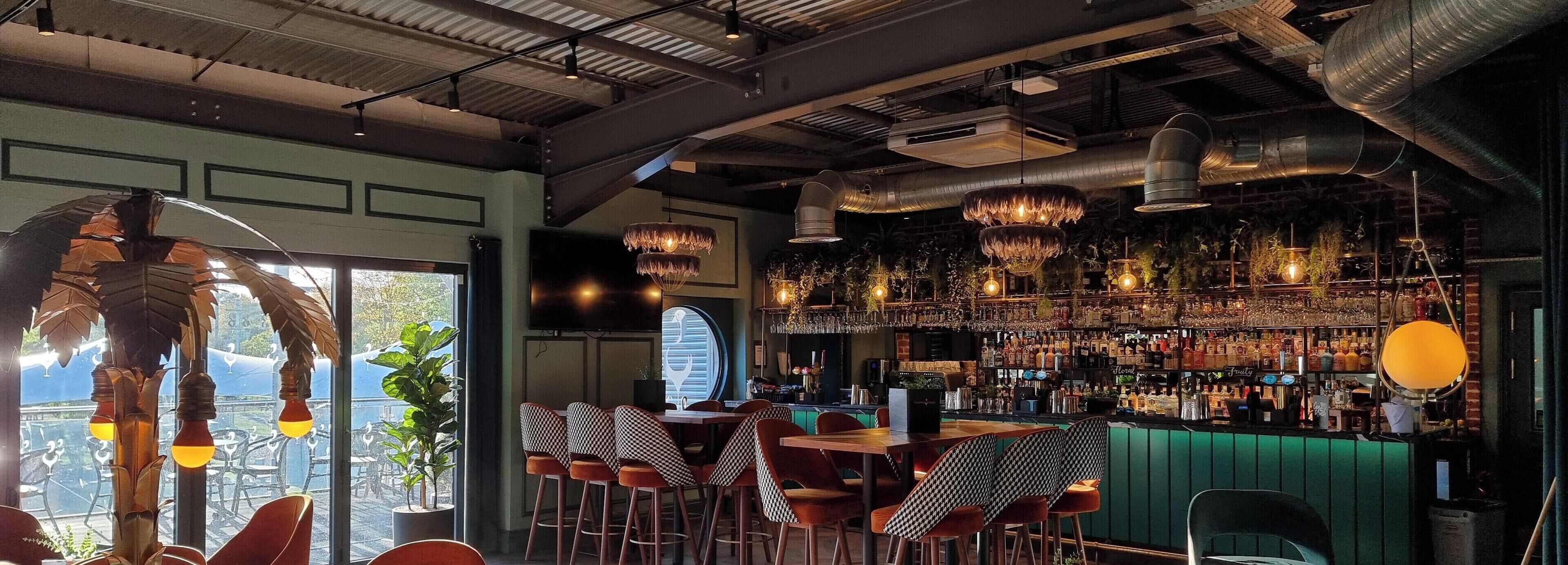 A bar area with a high ceiling, exposed ductwork, and a variety of hanging light fixtures. There are multiple seating areas with checkered tablecloths and bar stools.