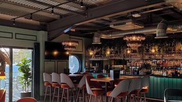 A bar area with a high ceiling, exposed ductwork, and a variety of hanging light fixtures. There are multiple seating areas with checkered tablecloths and bar stools.
