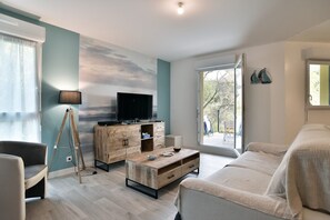 A modern living room with a wooden coffee table, a sofa, a TV on a wooden cabinet, and a large window with a view of the outdoors. - Chez Josse - A 10 min de St-Cast (Matignon)