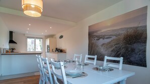 Un comedor con una mesa blanca dispuesta para seis personas, rodeada de sillas blancas, un cuadro grande en la pared y una cocina al fondo.