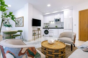 A modern living room with a round wooden coffee table, two armchairs, a sofa, a TV, and a kitchen in the background. - Genteel Home Uceda I (Cordoba)