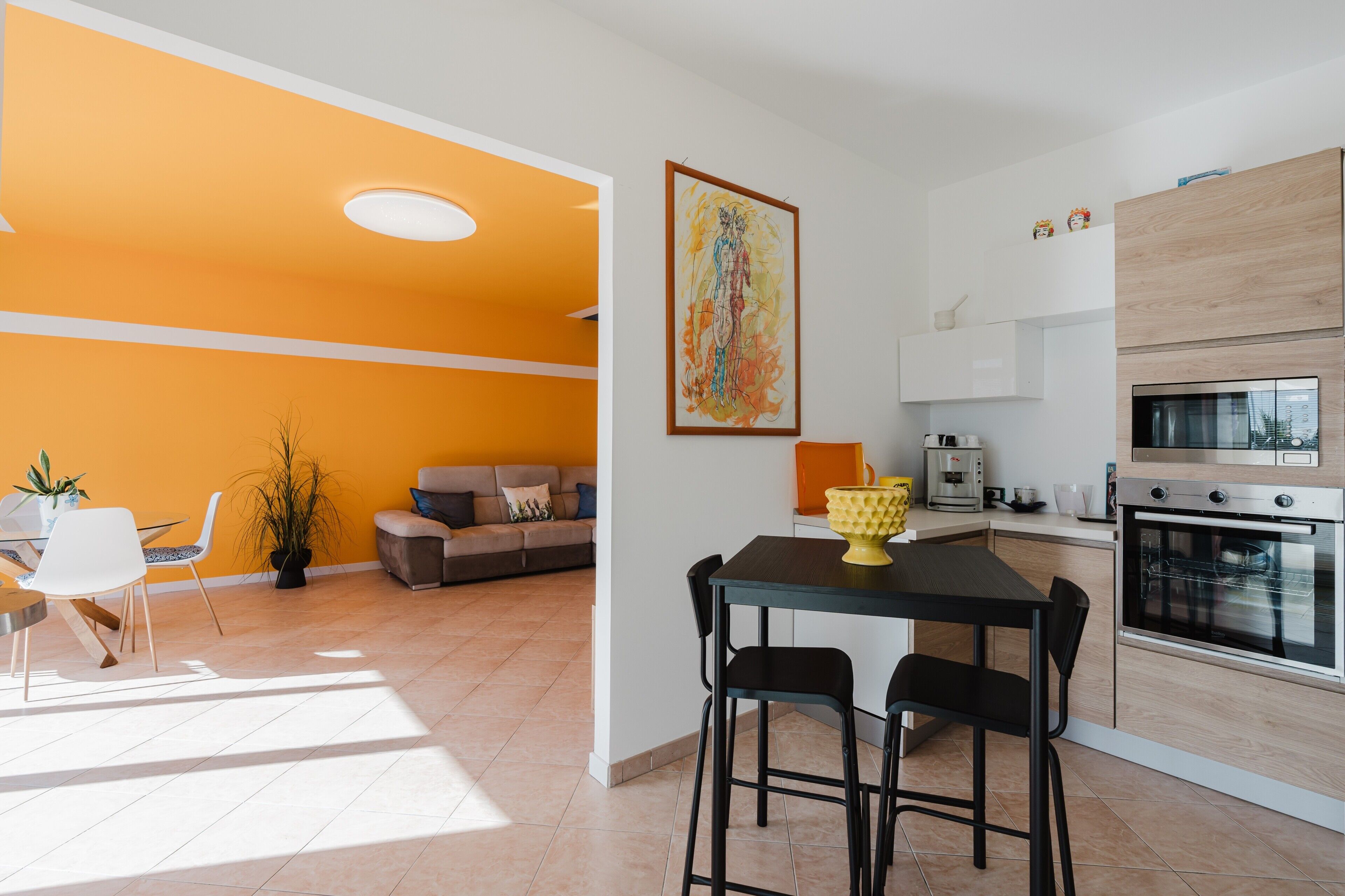 A modern kitchen with a built-in oven, a dining area with a table and chairs, and a living area with a sofa and a plant.