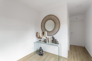 Un pasillo con una consola de vidrio que exhibe objetos decorativos, un espejo redondo en la pared y una puerta cerrada al final.