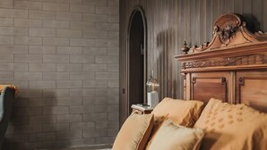 Una habitación con pared de ladrillo, cabecera de madera, cama con almohadas color beige y mesita de noche con lámpara.