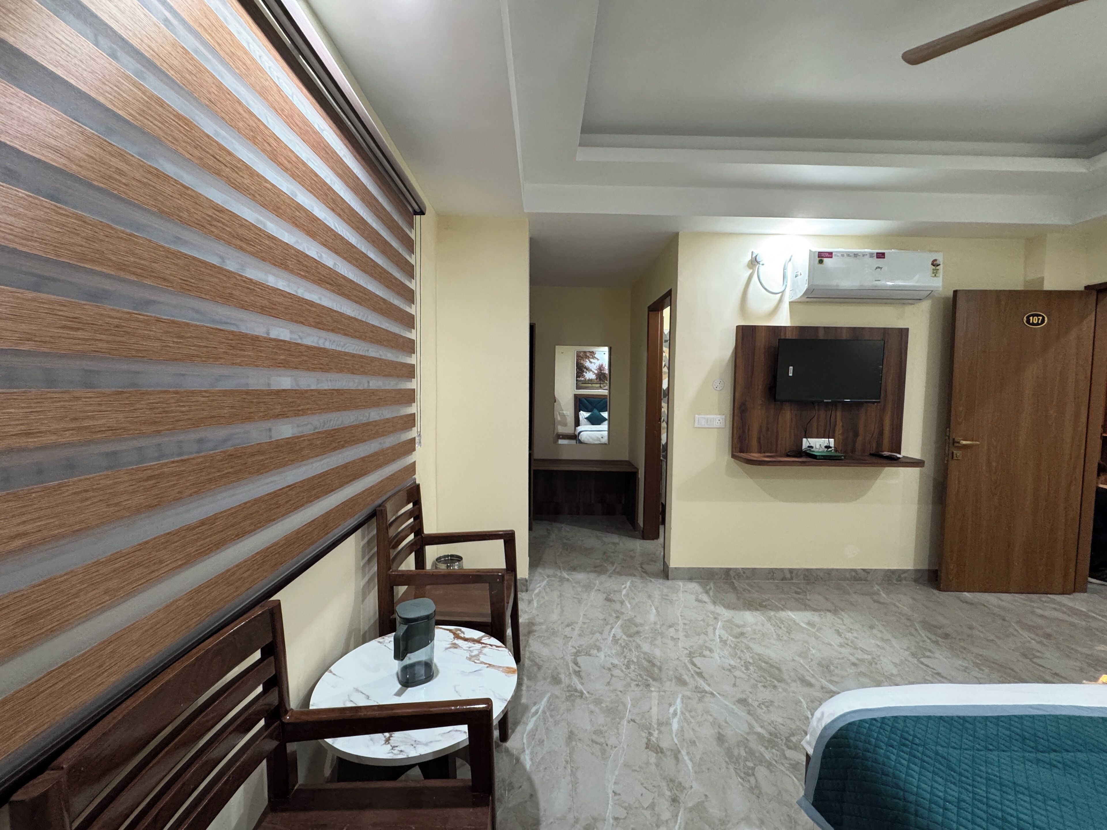 A hotel room with a wooden slatted wall, a round table with a chair, a bed, a television mounted on the wall, and a door with the number 107.