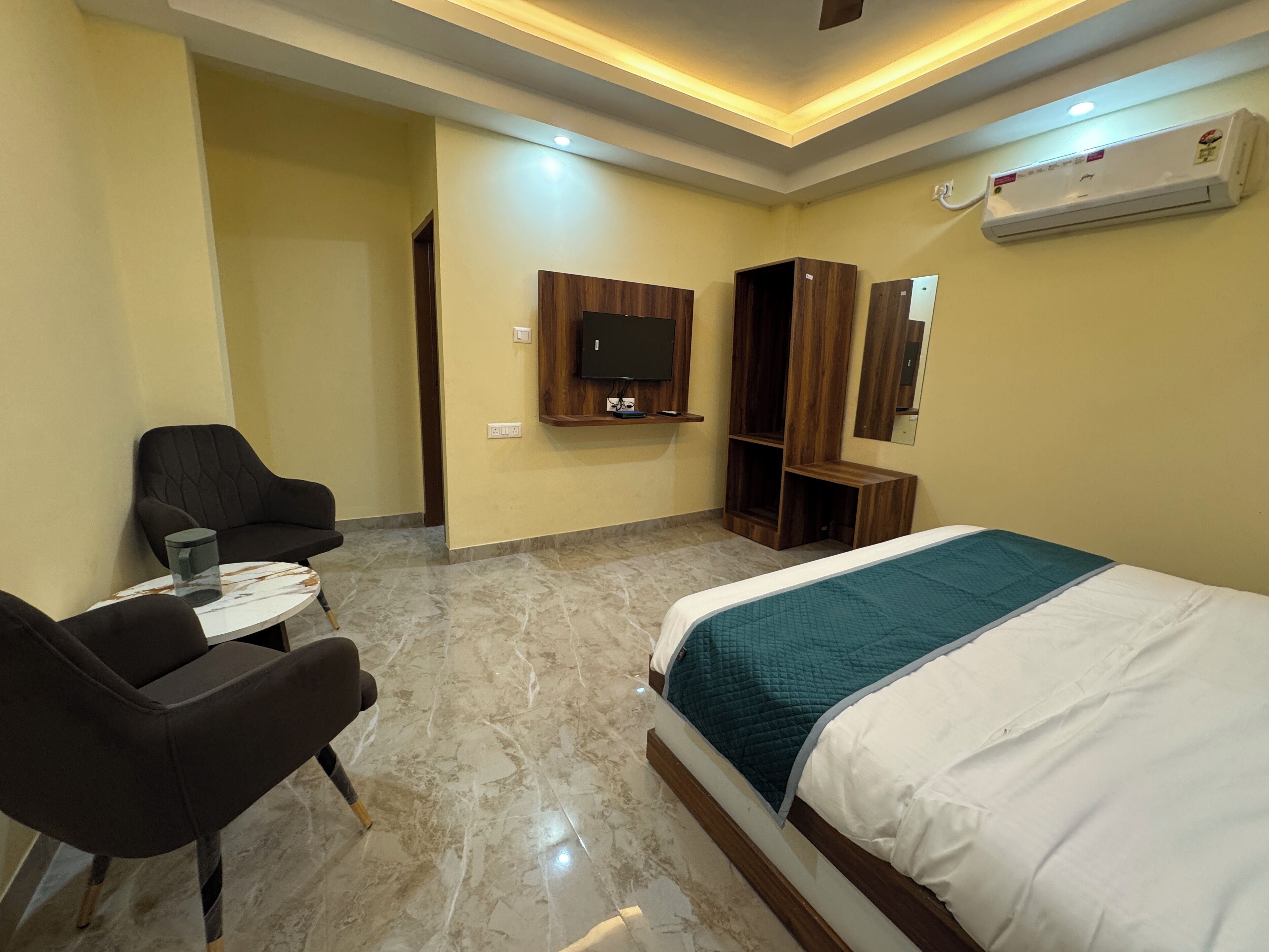 Une chambre d’hôtel comprenant un lit, un téléviseur fixé au mur, une armoire en bois, une petite table avec une chaise et un sol en marbre.