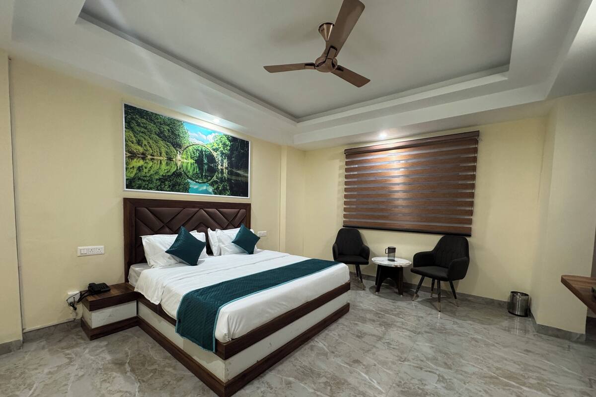 A hotel room with a large bed, two chairs, a small table, a ceiling fan, a window with blinds, and a painting on the wall.