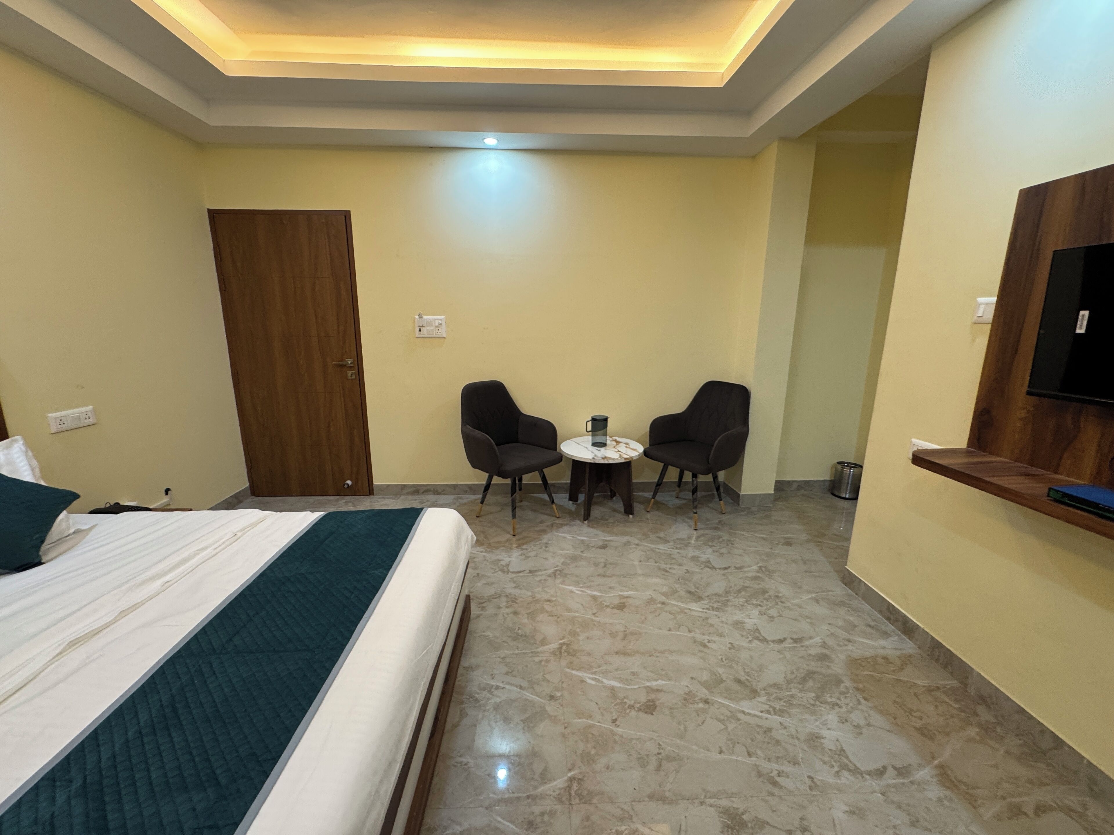 Habitación de hotel con cama, dos sillones, una mesa redonda pequeña, televisor montado en la pared y una puerta de madera.