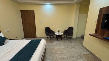 Habitación de hotel con cama, dos sillones, una mesa redonda pequeña, televisor montado en la pared y una puerta de madera.