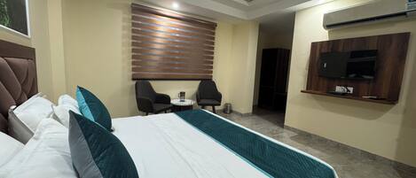 Habitación de hotel con una cama grande, dos sillones, una mesita, un ventilador de techo, un televisor montado en la pared y una ventana con persianas.
