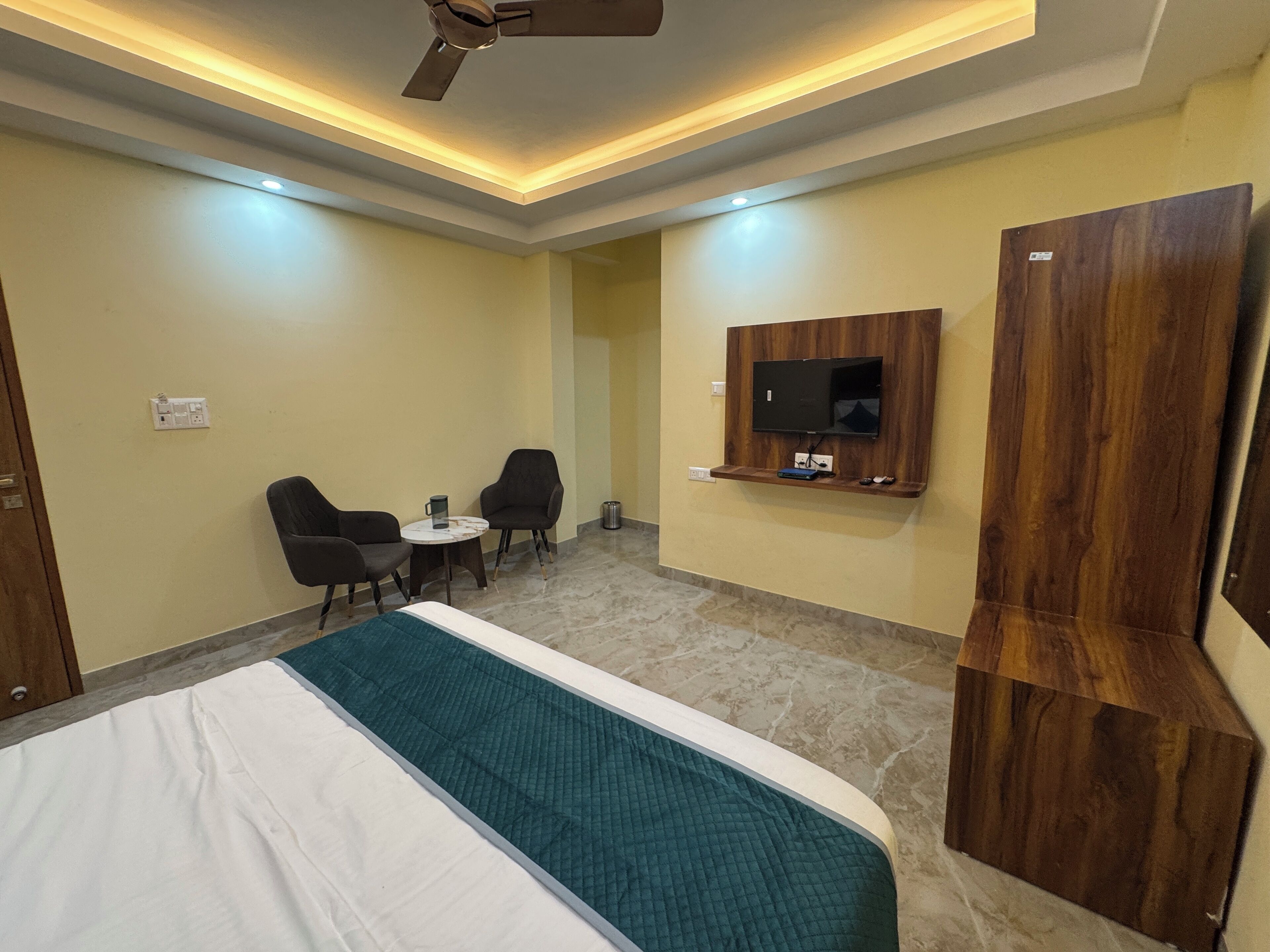 A hotel room with a bed, a TV mounted on the wall, a small table with two chairs, and a ceiling fan.