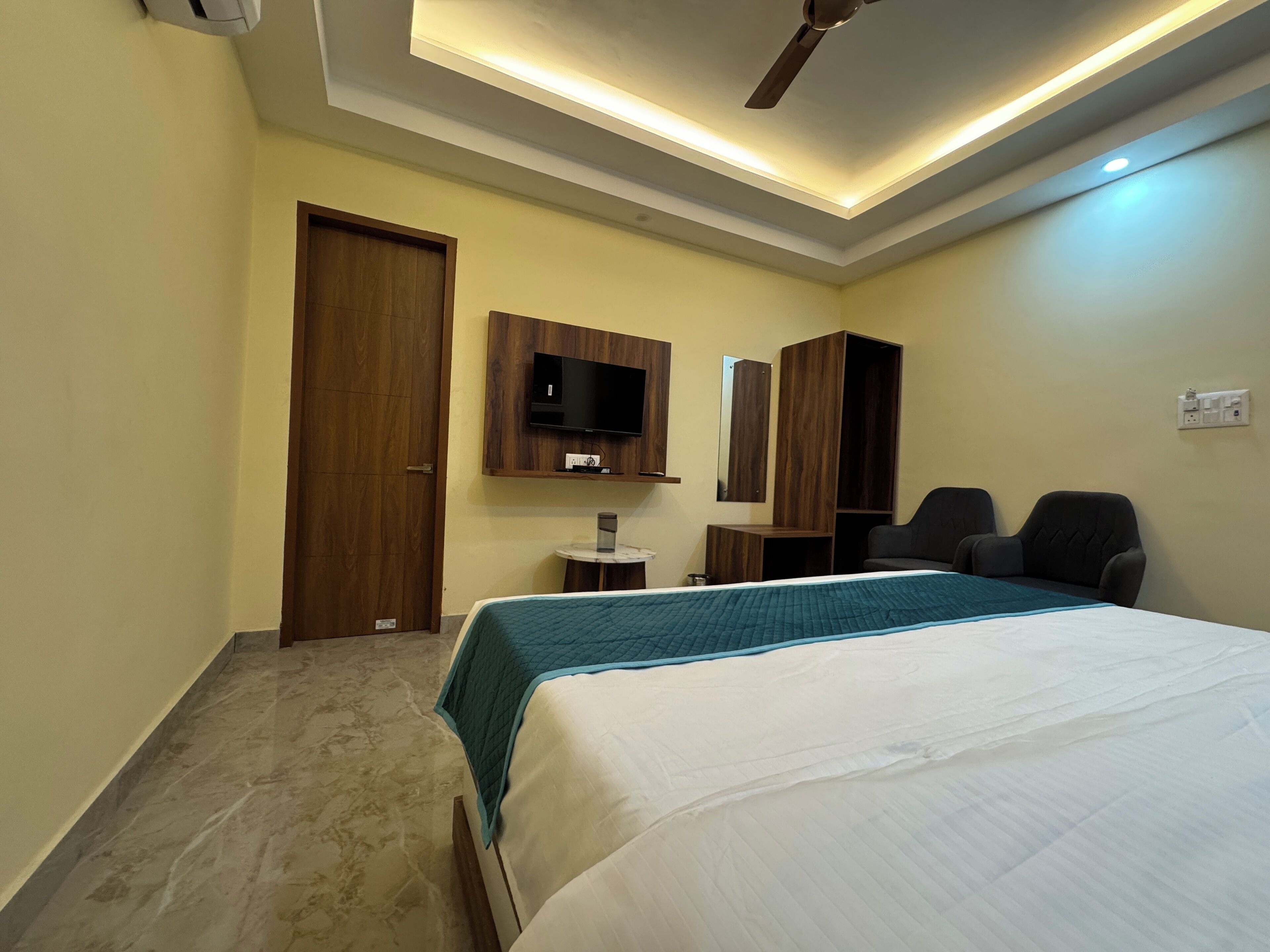 A hotel room with a bed, a TV mounted on the wall, a ceiling fan, and a small table with a lamp.