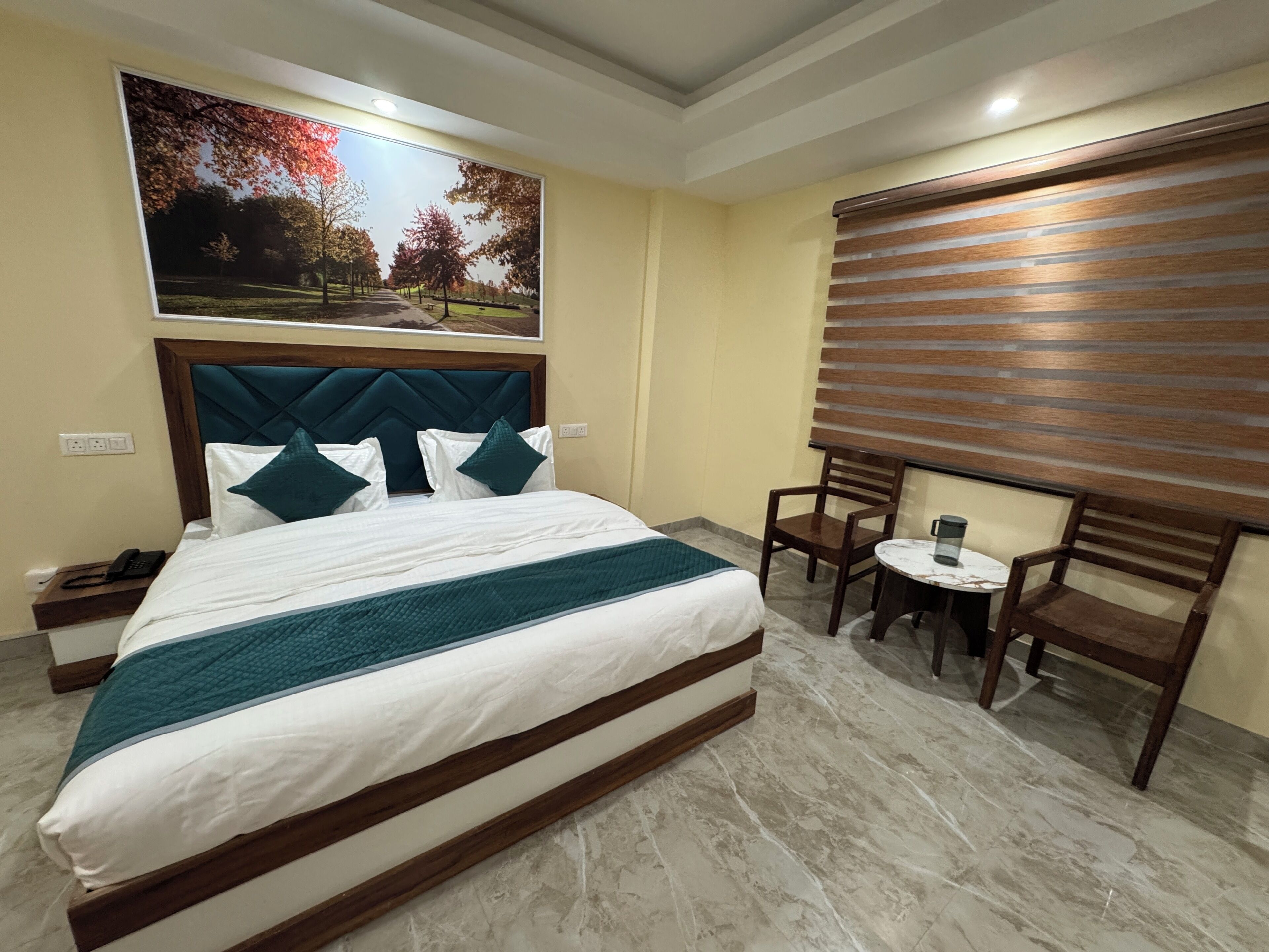 A hotel room with a large bed, a headboard, a painting of a park, a small table with chairs, and a window with blinds.