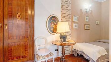 A room with a wooden door, a white chair, a round table with a lamp, a bed with white bedding, and a wall with framed pictures.