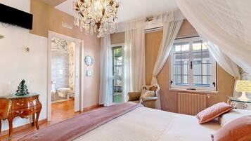 A bedroom with a bed, a chandelier, a window with curtains, a radiator, and a wooden dresser.