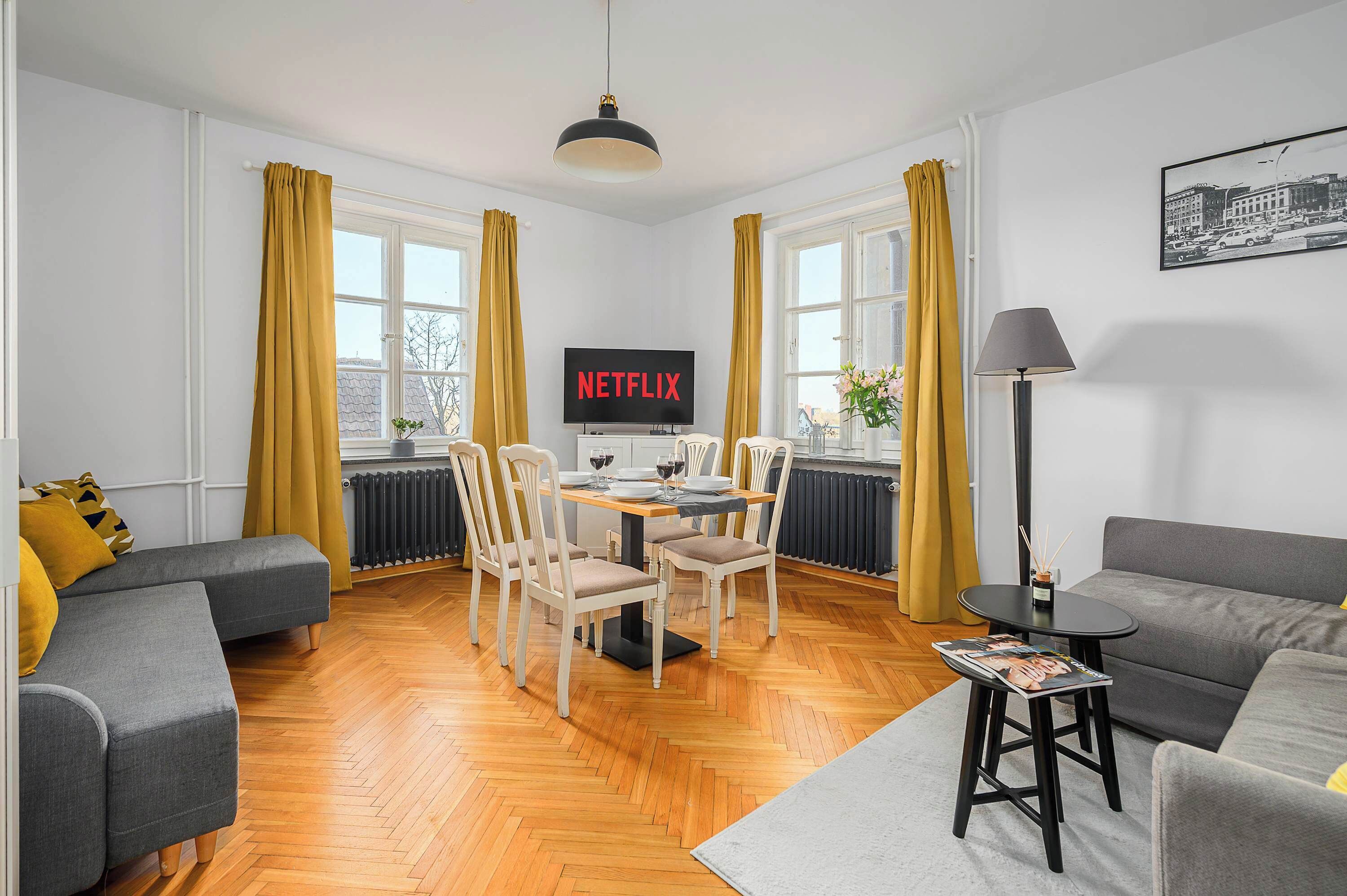 A modern living room with a dining area, featuring a flat-screen TV, wooden flooring, and a sofa.