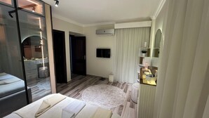 Ett hotellrum med en säng, en tv, en spegel, ett litet bord, ett sängbord, en nattduksbord, en lampa, ett fönster och en dörr.