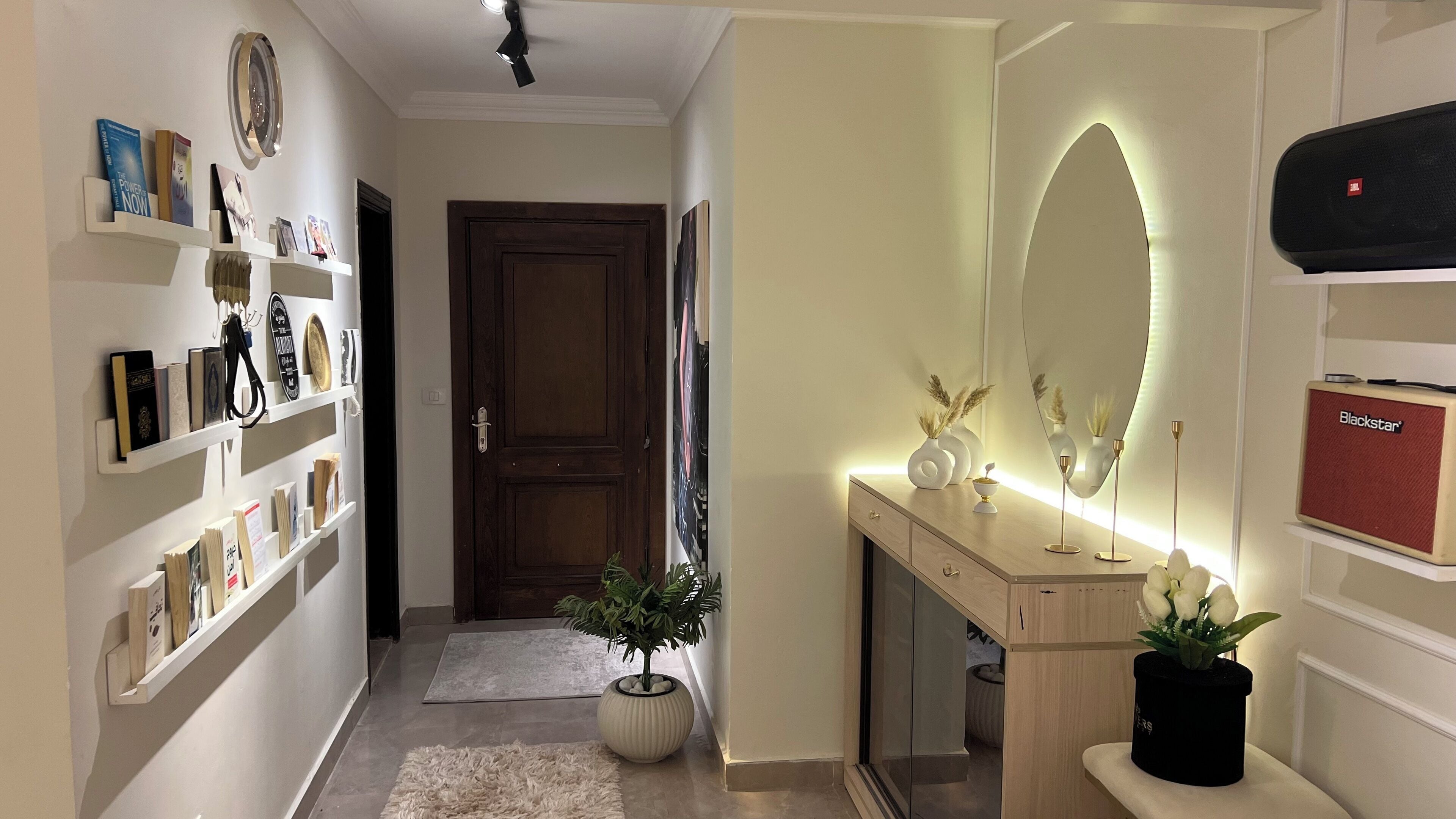 A hallway with a wooden door, a mirror with a light, and a Blackstar speaker on a shelf.
