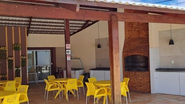 Une terrasse extérieure avec des chaises et des tables en plastique jaune, un four en briques et un espace couvert doté d’un toit en tuiles.