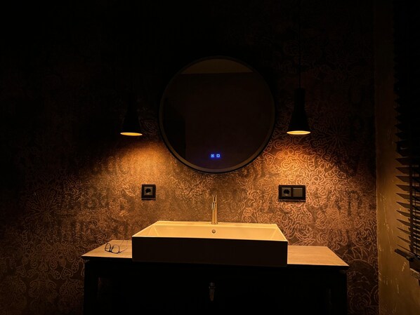 Une salle de bain avec un lavabo rectangulaire, deux appliques murales et un miroir rond.