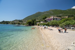 A beach with clear water, a dock, and buildings in the background. - Odysseia Apartment 1 - 2 Min Walk to Beach, 4 Adults, Terrace, Wi-Fi & AC (Nikiana)