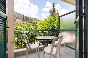 Un balcon avec une table ronde et quatre chaises blanches, donnant sur un jardin luxuriant et des collines lointaines.