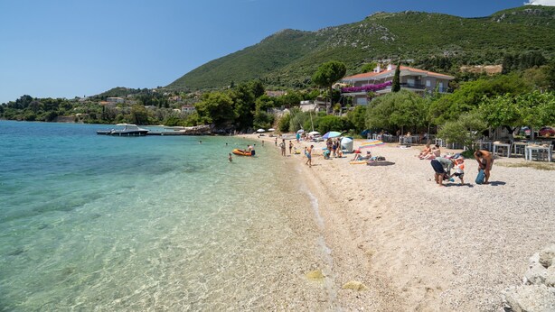 A beach with clear water, a dock, and buildings in the background. - Odysseia Apartment 4 - 2 Min Walk to Beach, 3 Adults, Terrace, Wi-Fi & AC (Nikiana)