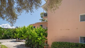 A building with a balcony, a paved area, and a tree. - Odysseia Apartment 4 - 2 Min Walk to Beach, 3 Adults, Terrace, Wi-Fi & AC (Nikiana)