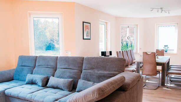 Una sala de estar moderna con un sofá seccional gris, un comedor con mesa y sillas, y ventanas amplias con vistas a árboles.