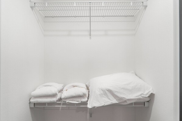 Ein weißer Kleiderschrank mit einem Metallregal, das gefaltete Handtücher und ein weißes Bett mit Decke beherbergt.