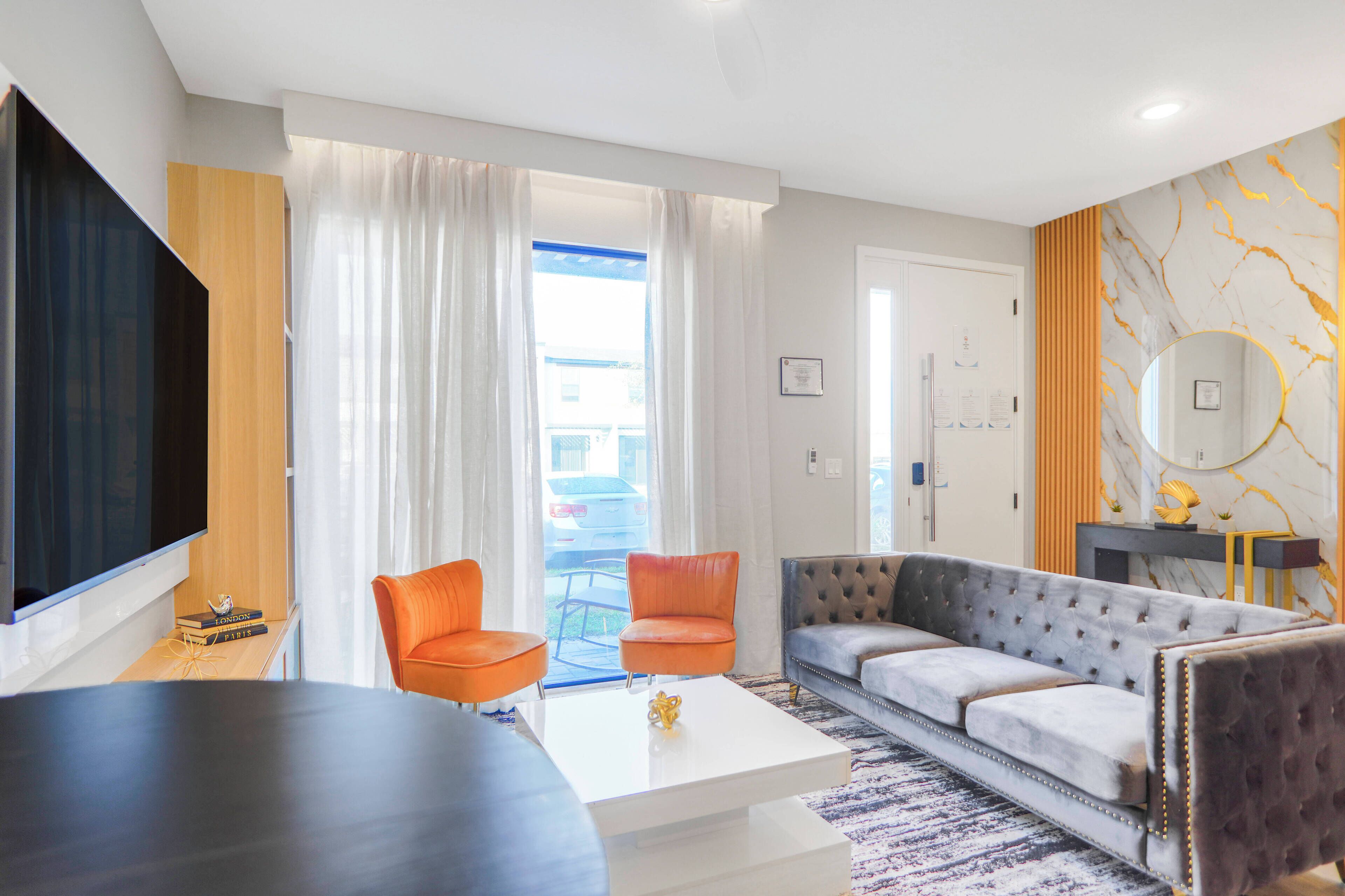 Un salon moderne avec un téléviseur à écran plat, deux fauteuils orange, un canapé gris et une table basse blanche.