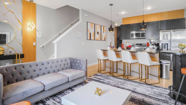 A modern living room with a grey sofa, a white coffee table, and a kitchen area with a dining set. - Luxury 3BR Home at The Hub Near Disney (Kissimmee)