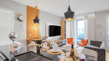 A modern living room with a flat-screen TV, a dining area with a table set for two, and a seating area with a sofa and chairs.