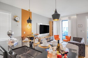 Uma sala de estar moderna com uma TV de tela plana, um espaço de refeições com mesa posta para dois e uma área de estar com sofá e cadeiras.