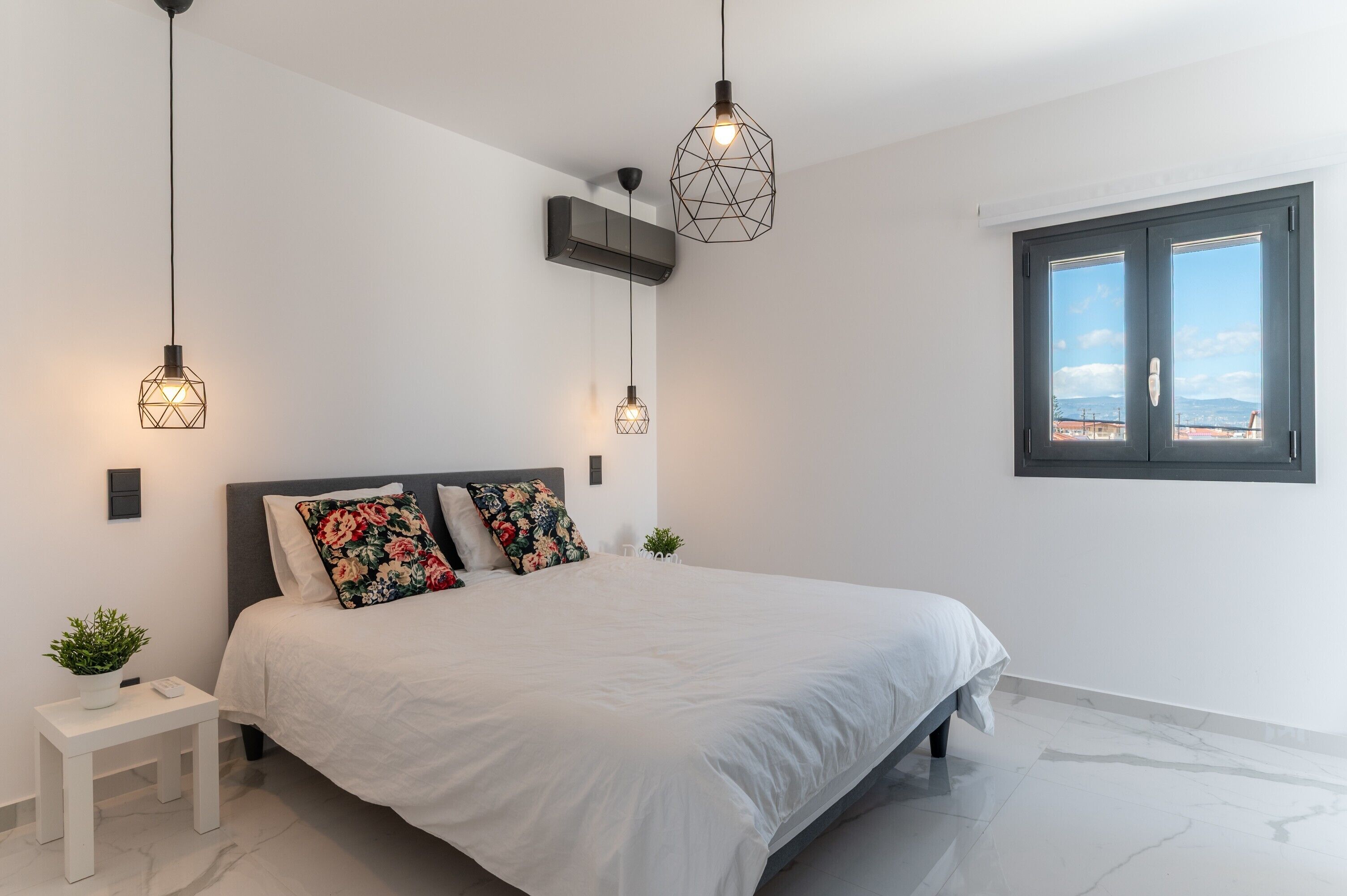 A modern bedroom with a bed, two pendant lights, a small table with a plant, and a window with a view.
