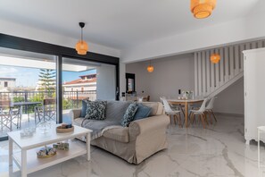 A modern living room with a sofa, dining table, and chairs, a staircase, and large windows.