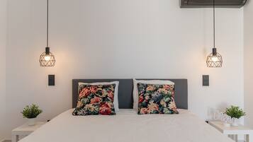 A bed with floral pillows, a white bedside table, a small plant, and a wall-mounted air conditioner.