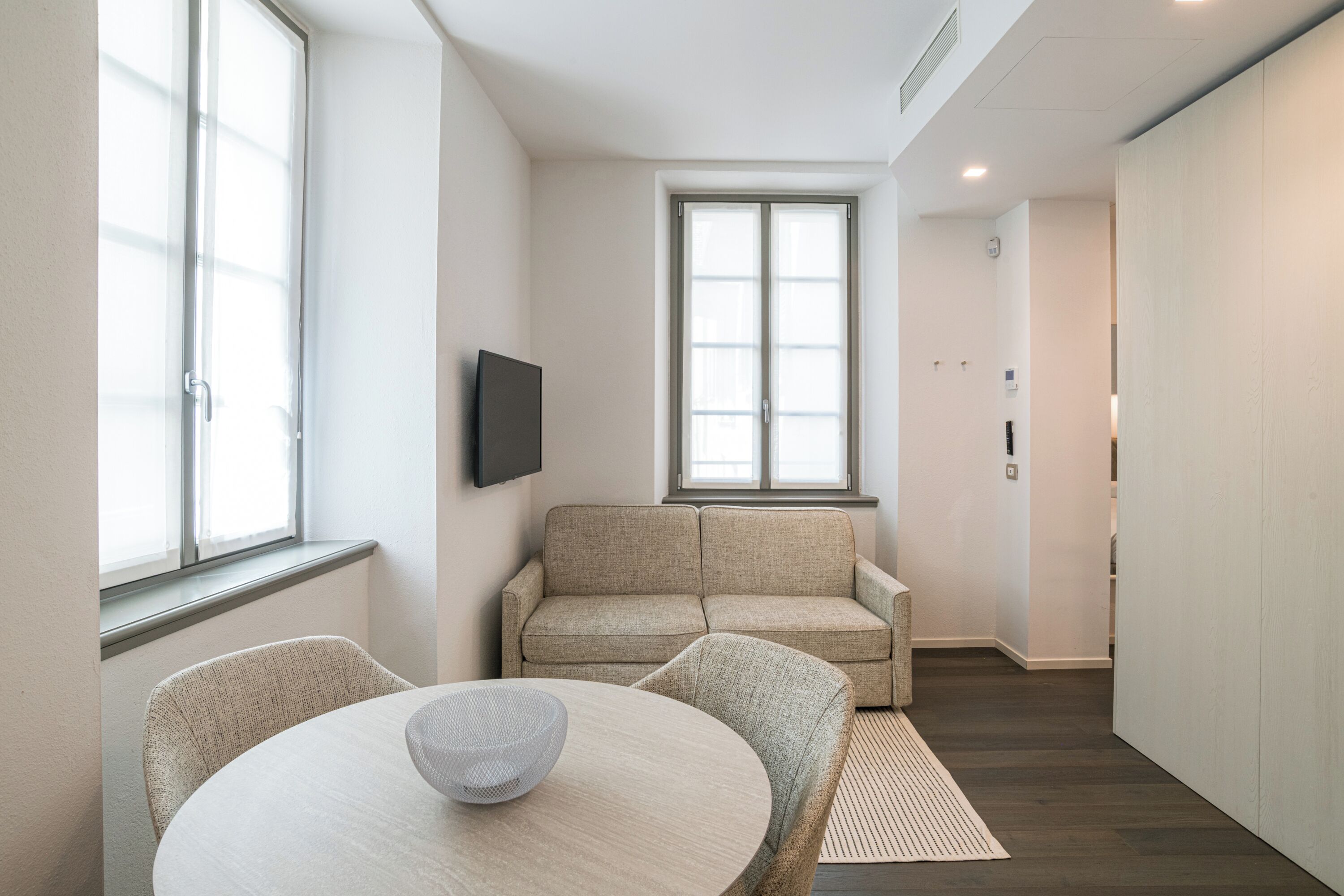 A modern living room with a sofa, a TV mounted on the wall, and a dining table with chairs.
