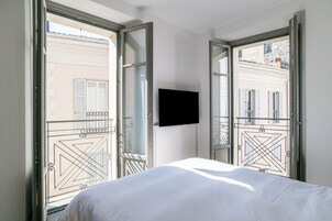 Habitación de hotel con cama, televisión montada en la pared y ventanas amplias con vistas a edificios.