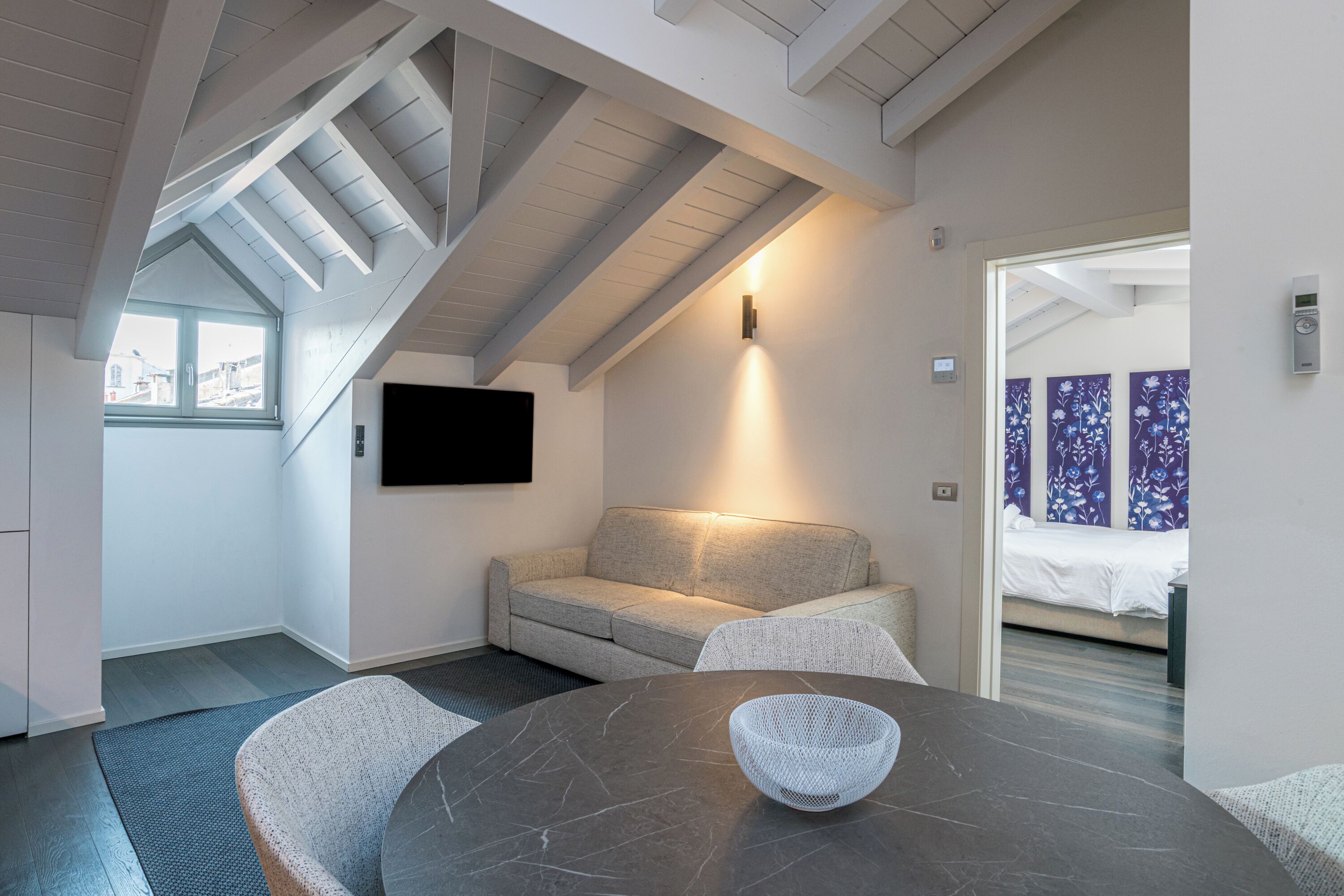 Quarto moderno com mesa de mármore escura, uma tigela trançada, sofá cinza e televisão na parede.