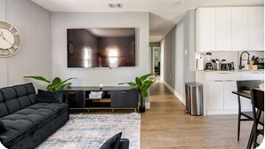 A modern living room with a black sofa, a TV mounted on the wall, and a kitchen area with white cabinets and a stainless steel sink. - MIAMI-close to downtown/Wynwood/mid-town/Brickell (Miami)