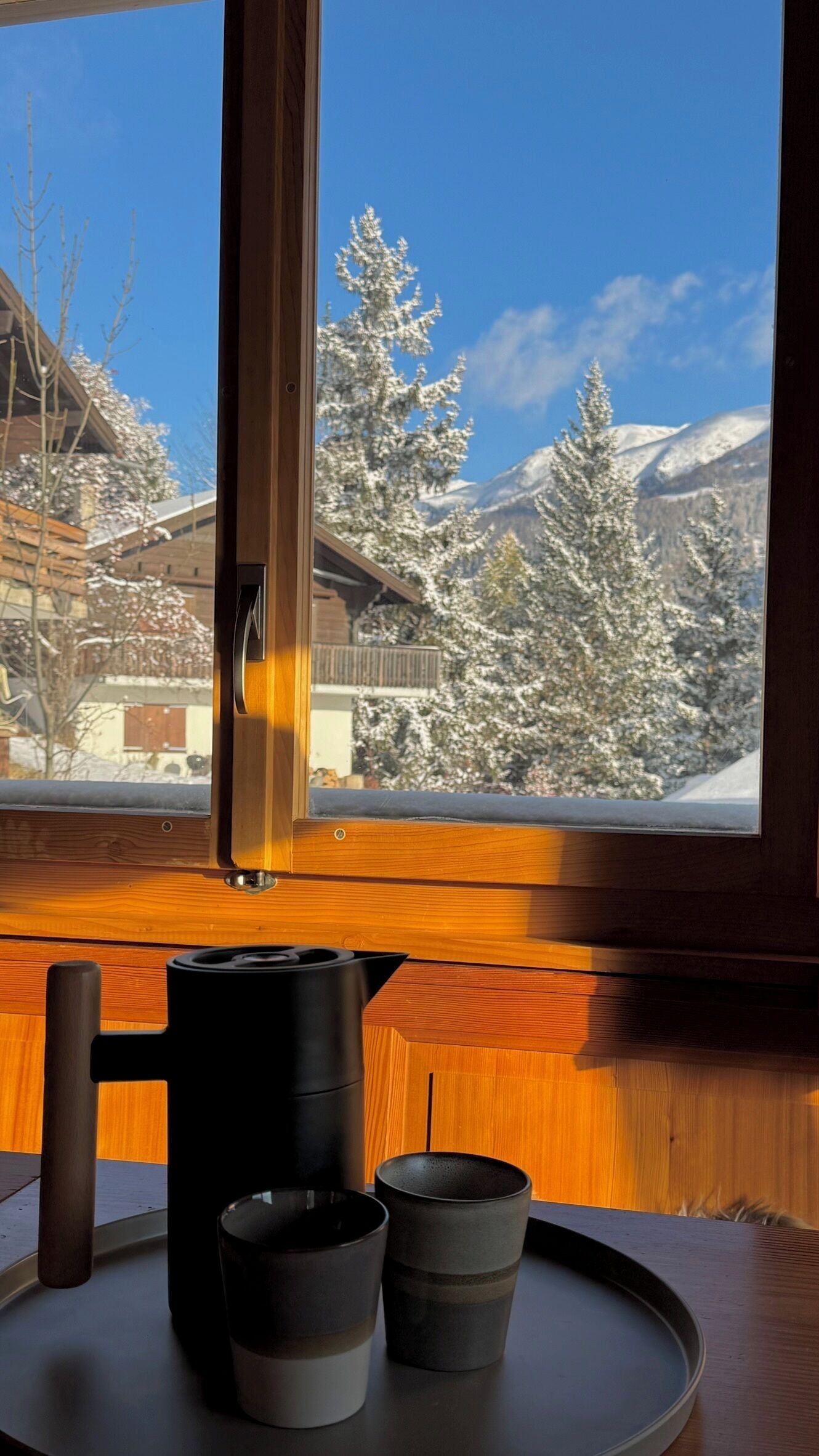 Ein Holztisch mit einer schwarzen Kaffeekanne und zwei Tassen, ein Fenster mit Blick auf schneebedeckte Bäume und Berge.