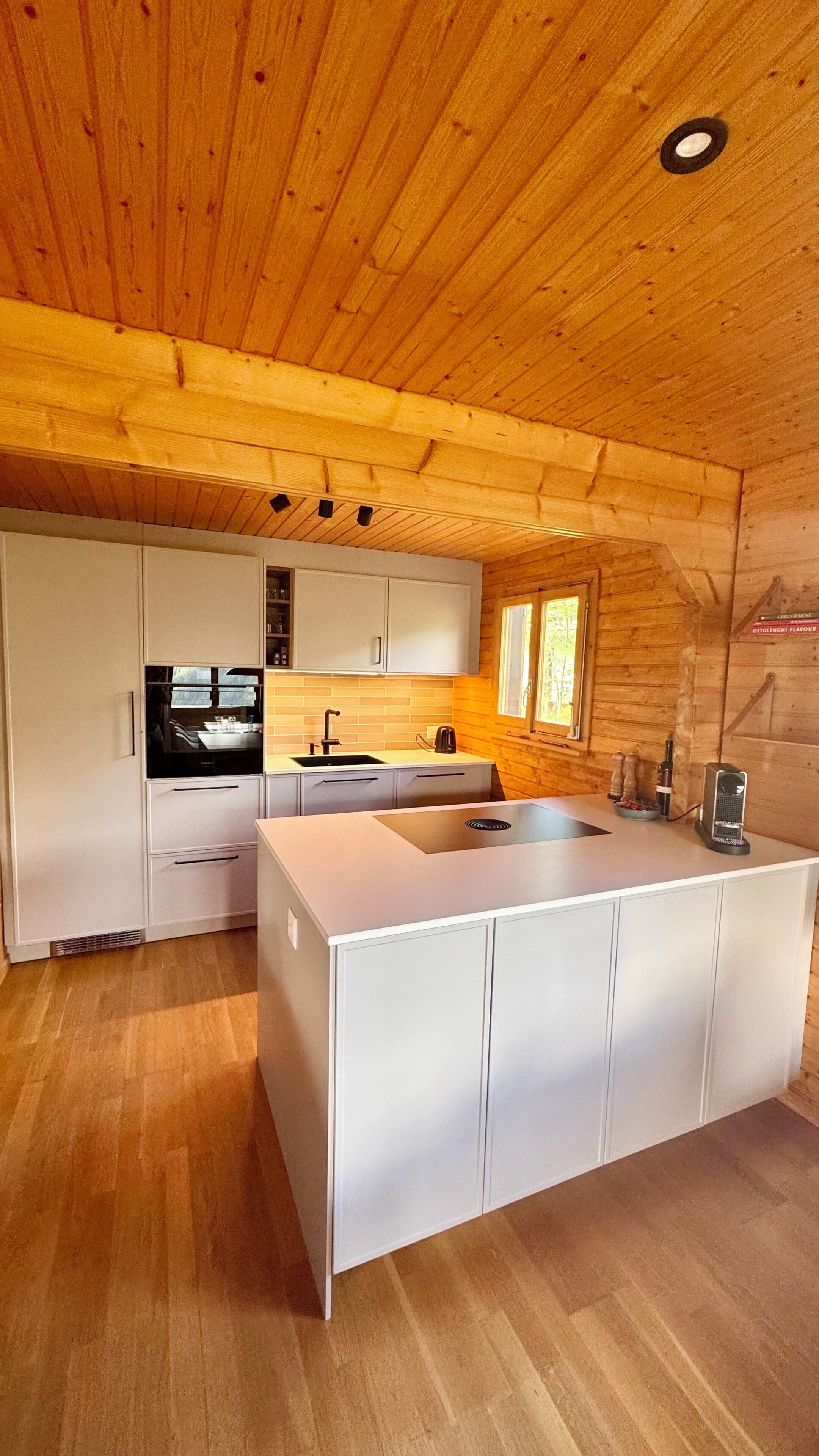 Une cuisine moderne avec des meubles blancs, un îlot central, une fenêtre et des poutres en bois au plafond.