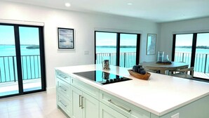 Een moderne keuken met een groot eiland, lichtblauwe kastjes en uitzicht op zee door grote ramen.