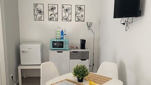 Un ambiente moderno e funzionale con angolo pranzo, caratterizzato da un tavolo bianco, quattro sedie bianche, una piccola pianta in vaso e un telecomando. Un forno a microonde e una macchina da caffè sono posizionati su un mobiletto bianco, mentre un televisore a schermo piatto è fissato alla parete.